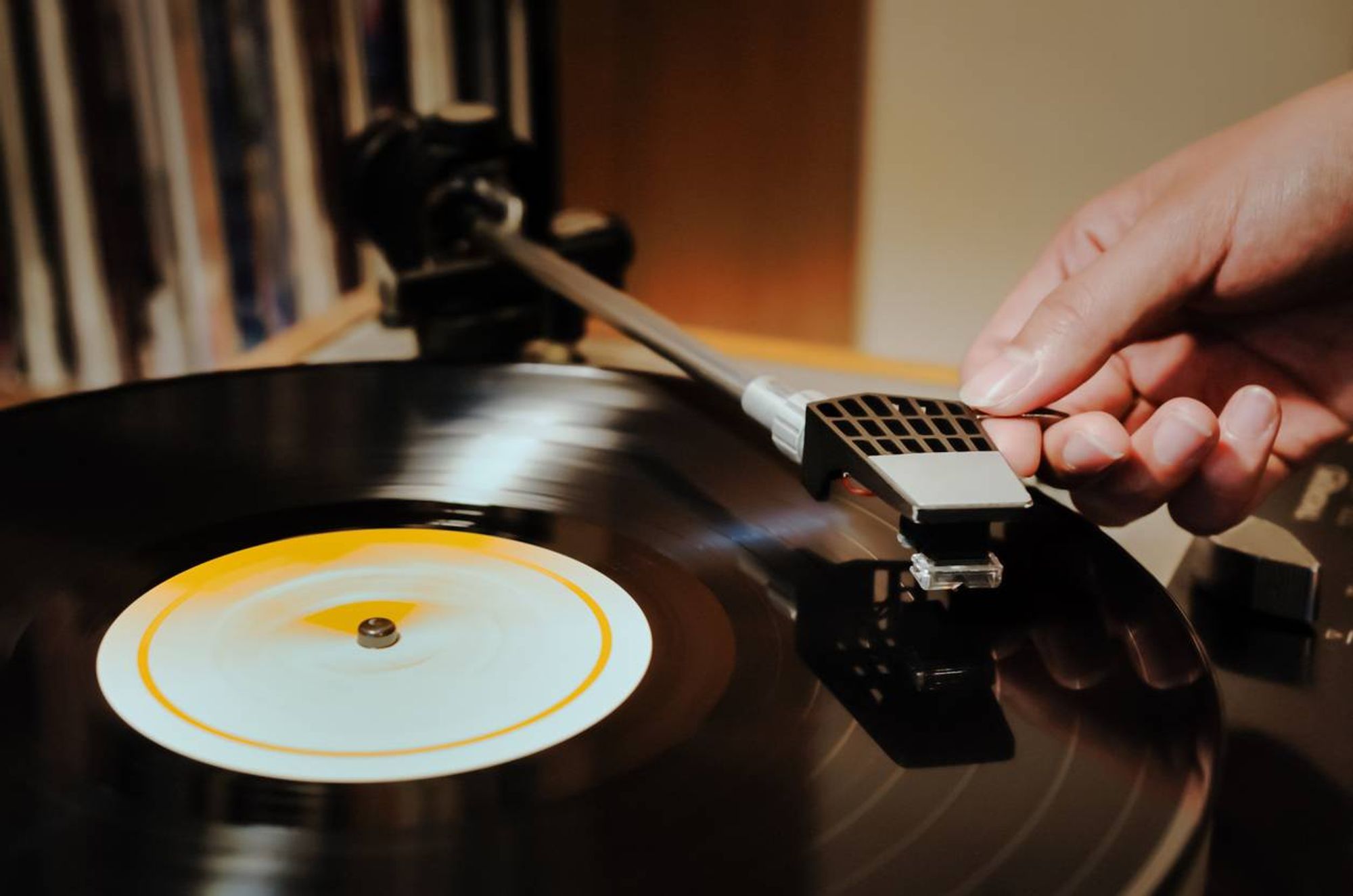 Close-up of a hand placing a needle on a spinning vinyl record on a turntable, with a blurred background of shelves.