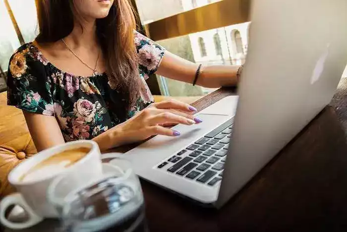 Woman typing on a laptop at a cafe table, with a floral top and purple nails. A cup of coffee and a glass of water are on the table.